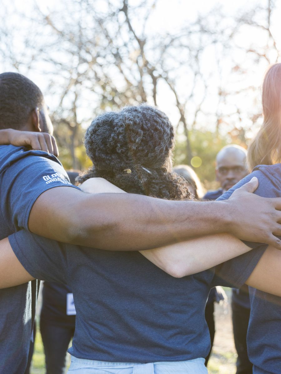 A group of people in blue shirts stand outdoors, arms around each other in a circle, facing away.