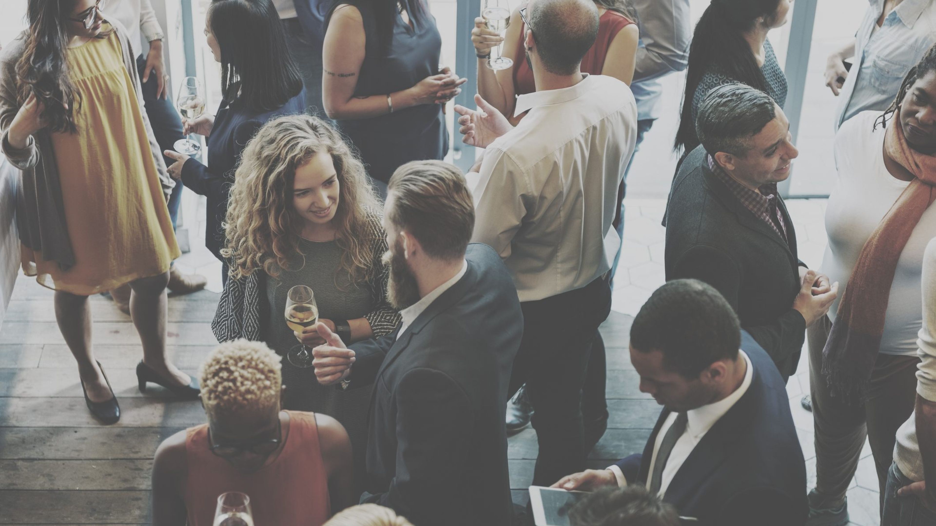 People at a social gathering, standing and talking while holding drinks in a bright indoor setting.