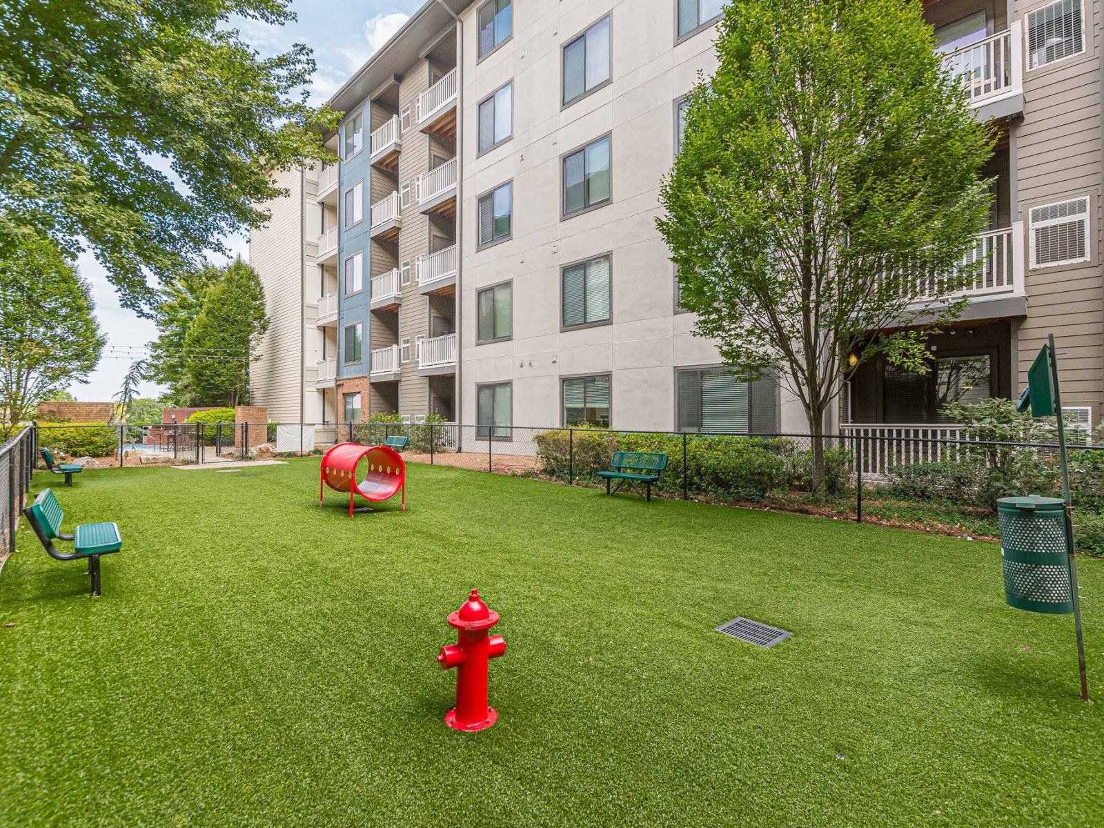 Apex West Midtown fenced grassy dog park area with benches, play tunnels, and a red fire hydrant beside an apartment building.