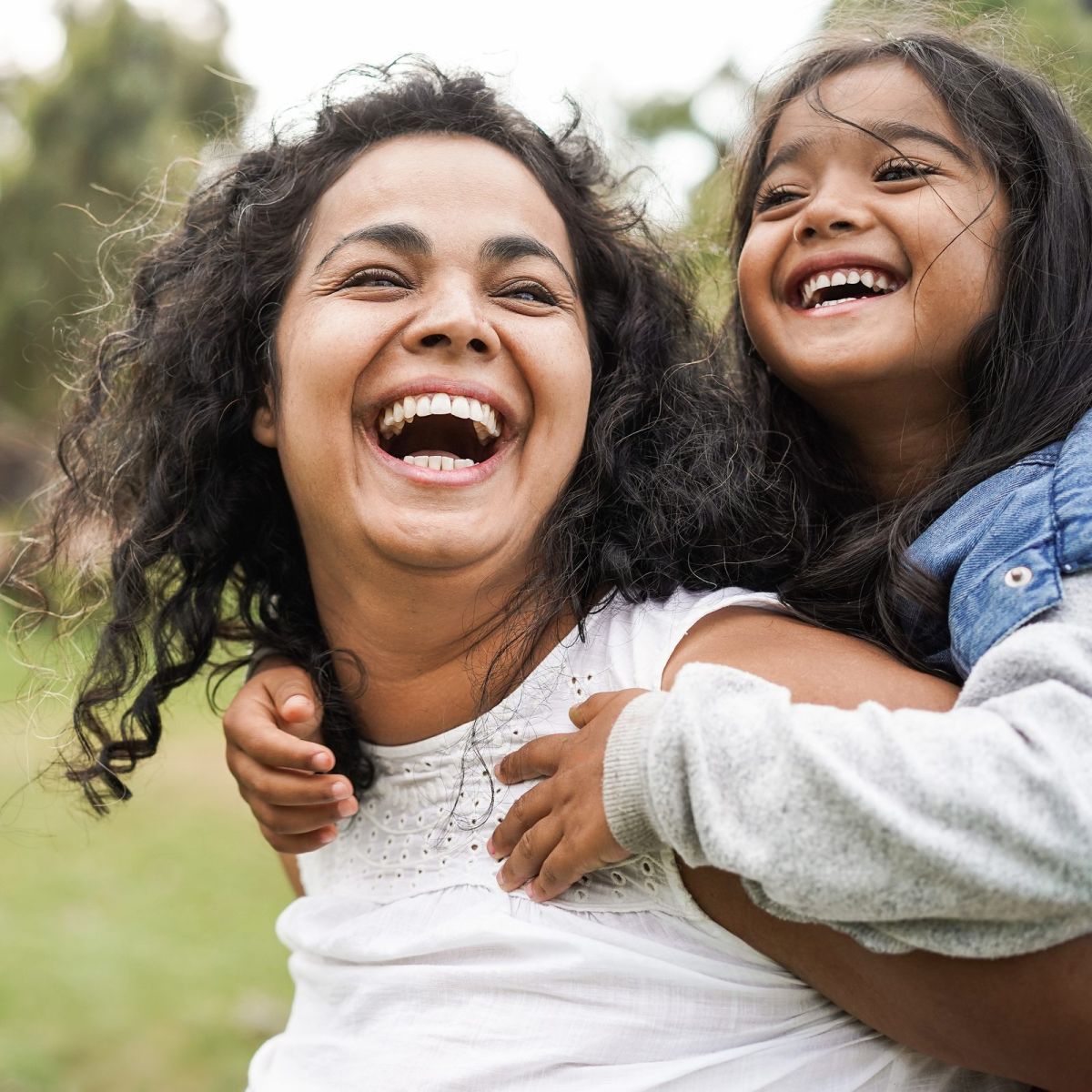 Woman and child smiling and laughing together outdoors in a park, with greenery in the background.