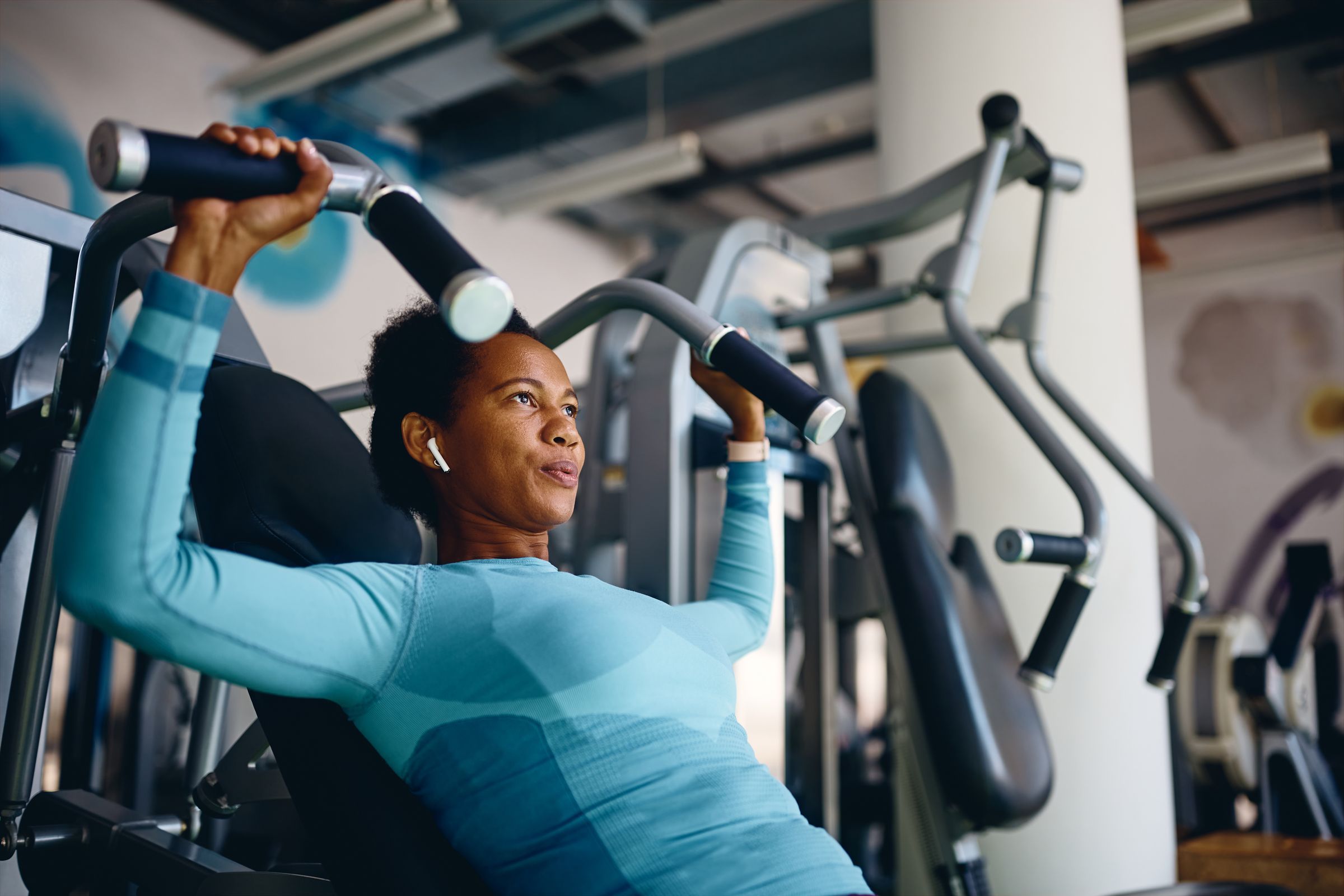 Woman in blue workout clothes using a chest press machine at the gym, wearing wireless earbuds.