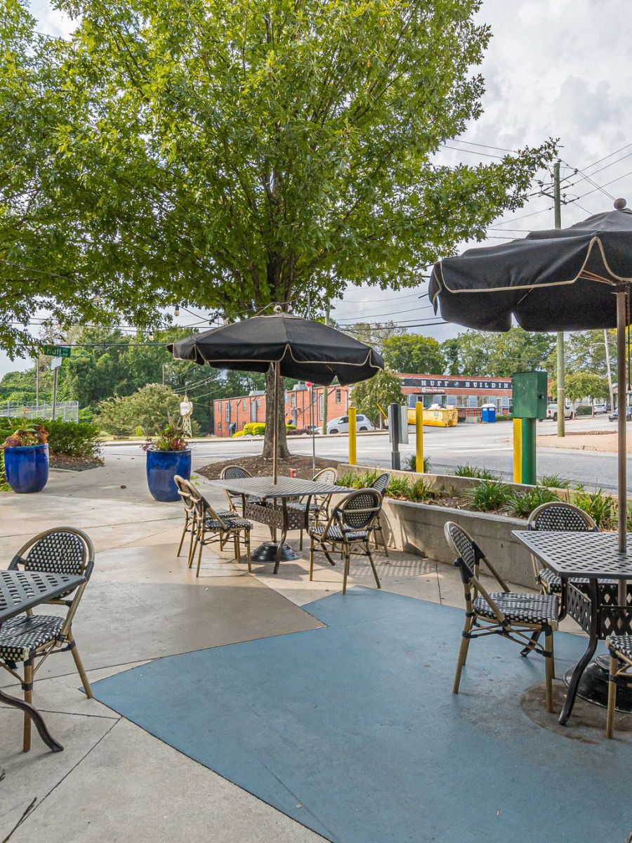 Outdoor café seating area with black tables, chairs, and umbrellas, surrounded by trees and street view.