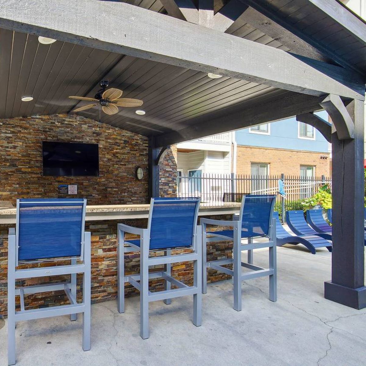 Apex West Midtown outdoor bar area with stone counter, blue chairs, TV, ceiling fan, and shaded pergola.