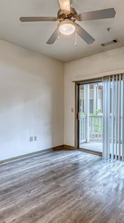 Apex West Midtown apartment room with wood flooring, ceiling fan, and sliding glass door with vertical blinds leading to a patio.