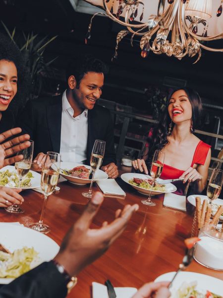 Six people dressed nicely laugh and talk while dining together at a restaurant table with food and drinks.
