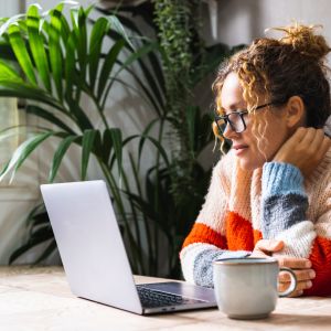 Woman with glasses using a laptop at a table, with a mug and a plant in the background.