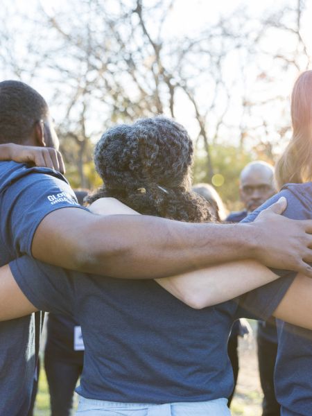 A group of people in blue shirts stand outdoors, arms around each other in a circle, facing away.