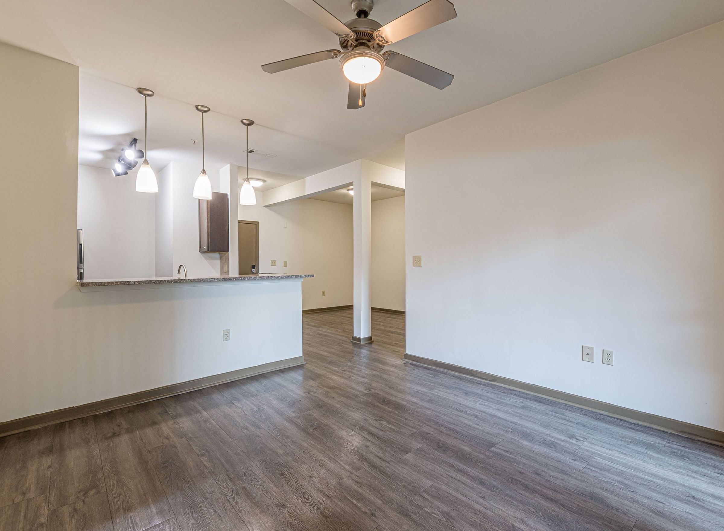 Empty modern room with wood floors, ceiling fan, and kitchen area with hanging pendant lights at Apex West Midtown.