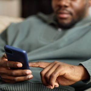 A person in a green sweater sits on a couch, using a smartphone with both hands.