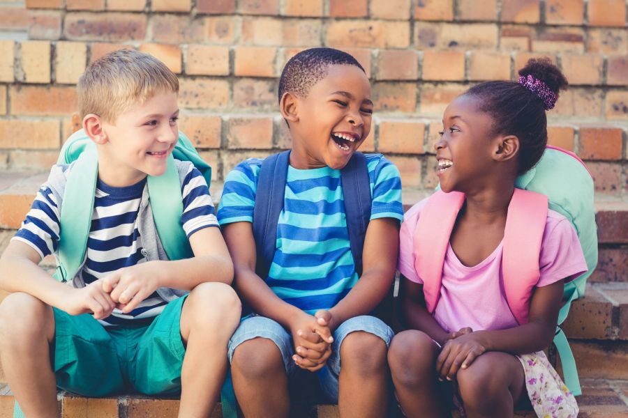 Three children with backpacks sit on steps, smiling and laughing together.
