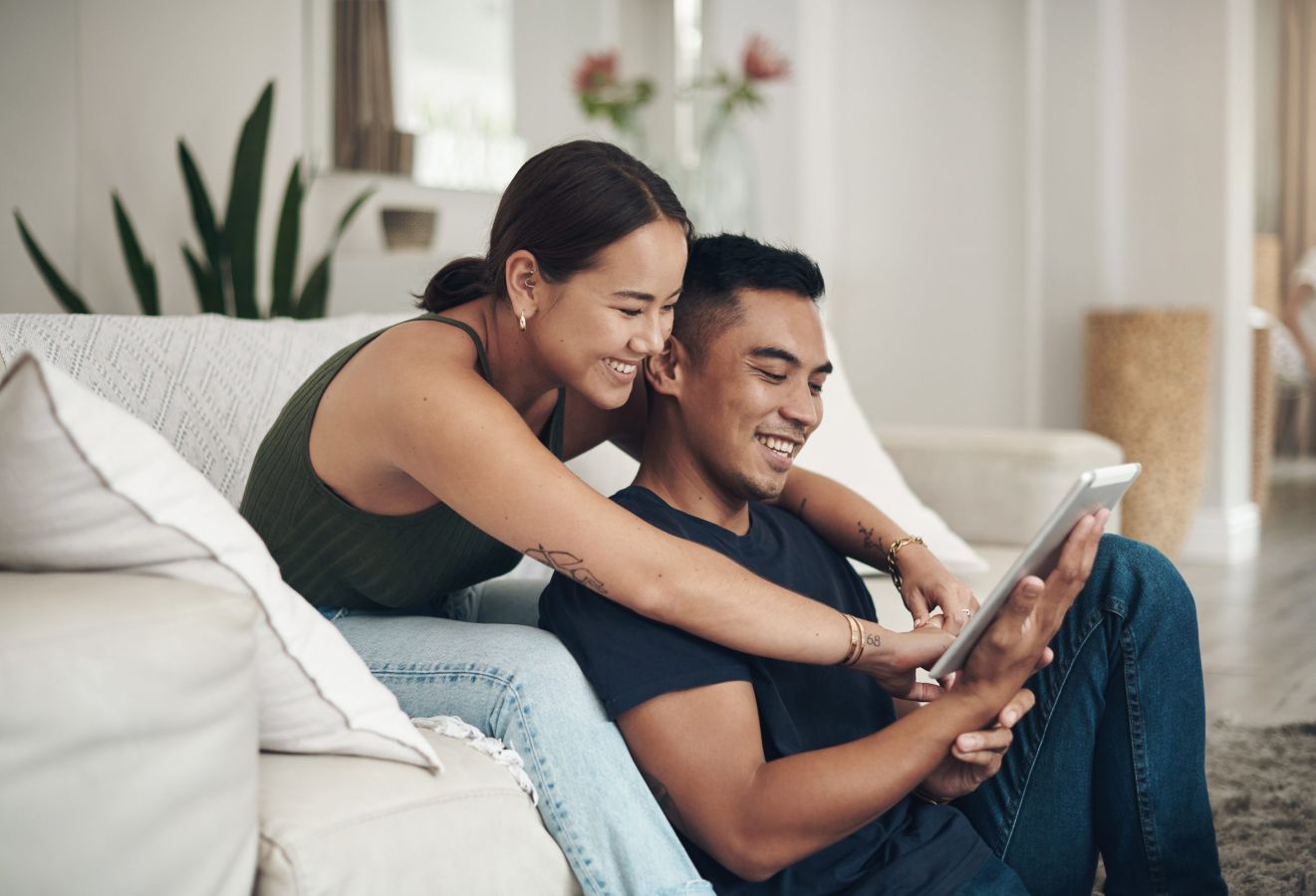 Smiling couple sitting on the floor, looking at a tablet together in a cozy living room.