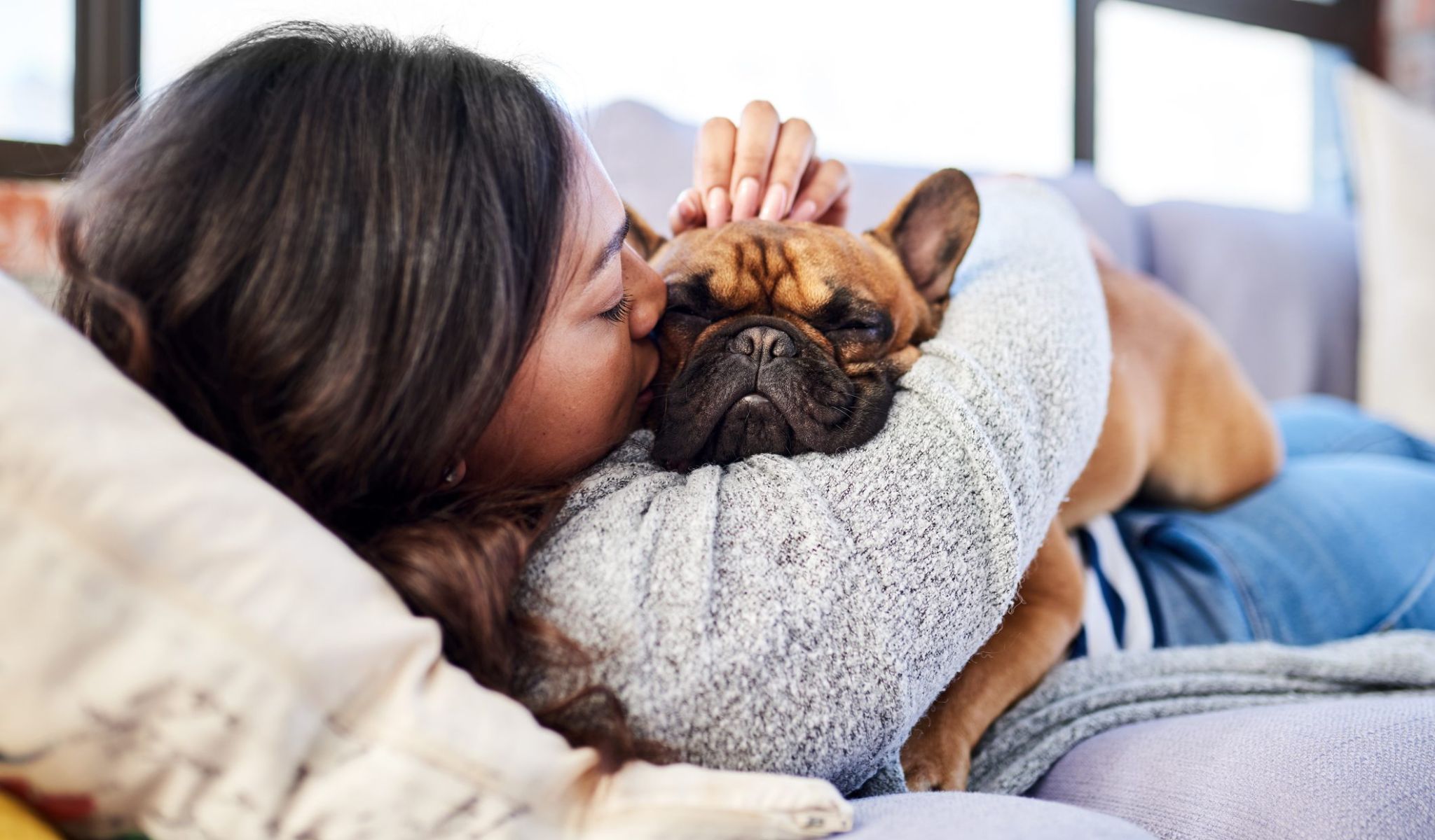 Woman lying on a couch hugging and kissing a brown French bulldog, both looking relaxed and cozy.