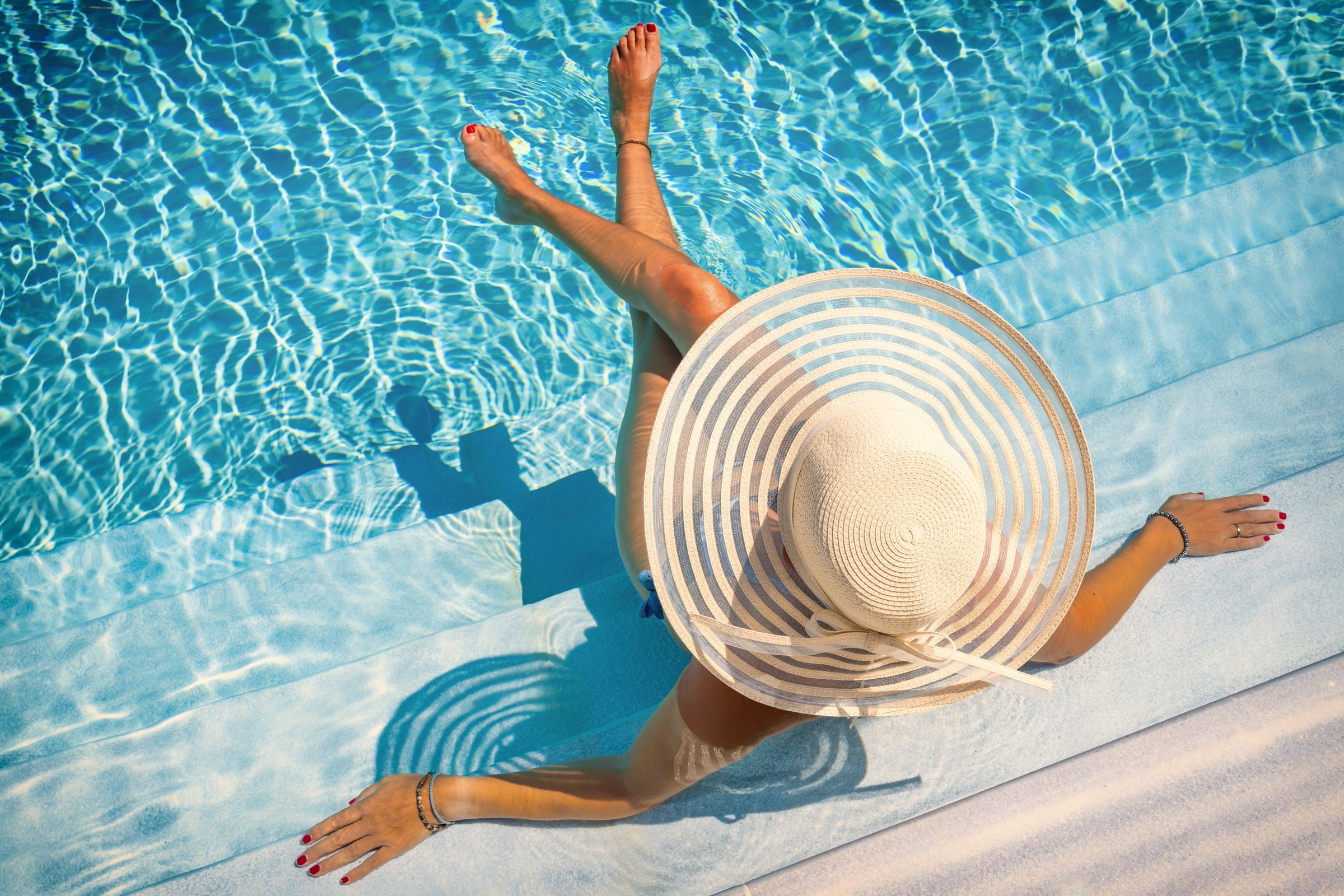 Woman in a striped sunhat sits on pool steps with feet in water, enjoying a sunny day.