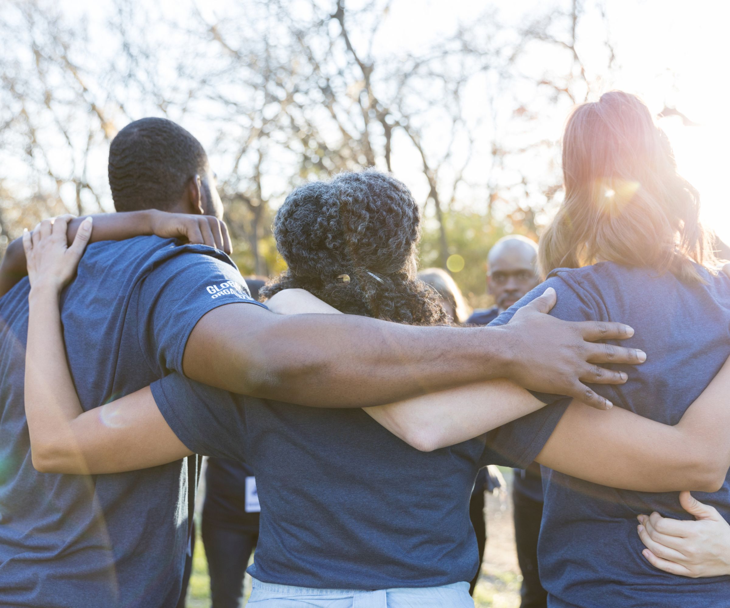 A group of people in blue shirts stand outdoors, arms around each other in a circle, facing away.