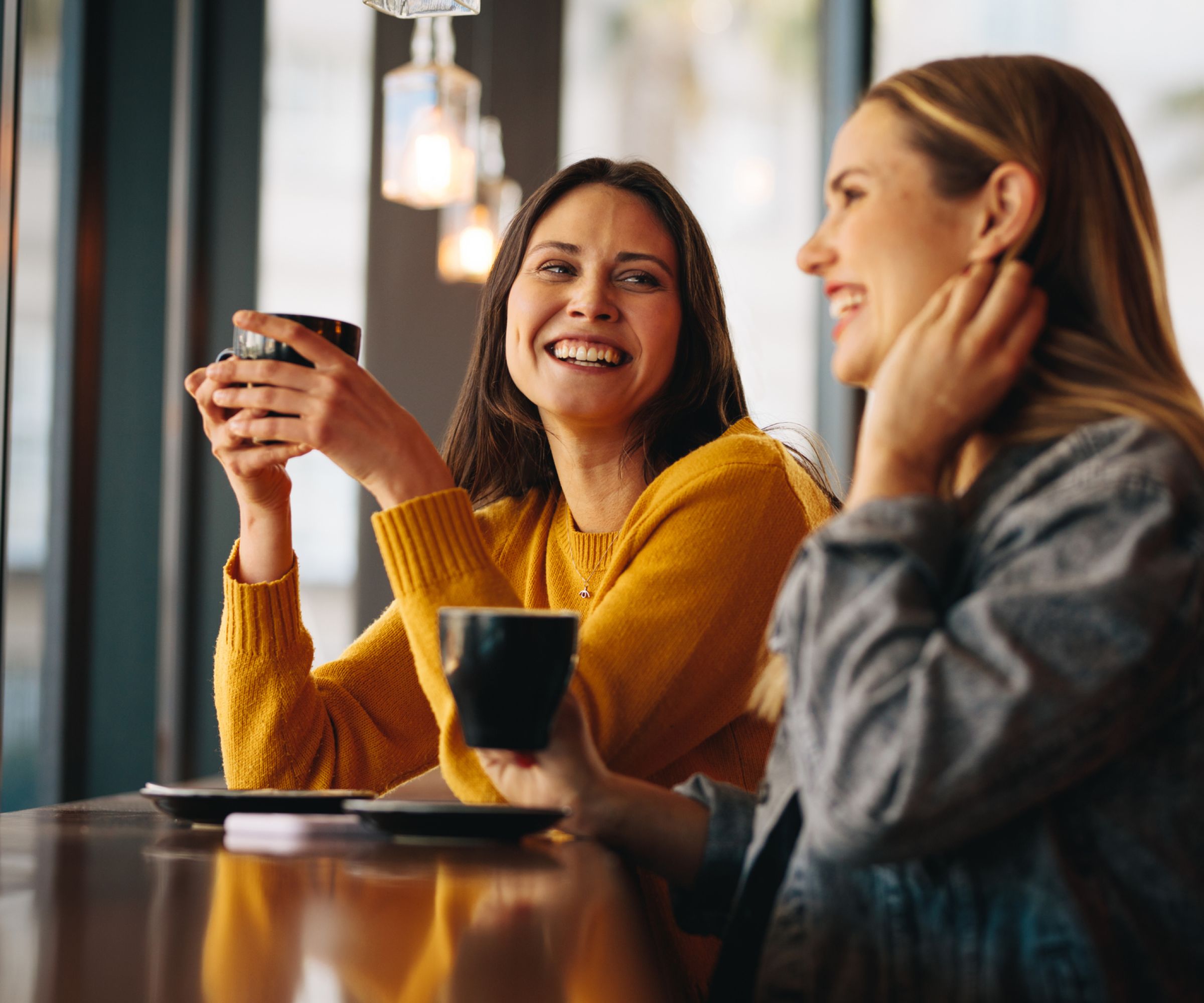 Two women smiling and chatting while holding coffee cups at a café table by the window.