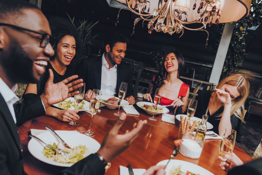 Six people dressed nicely laugh and talk while dining together at a restaurant table with food and drinks.