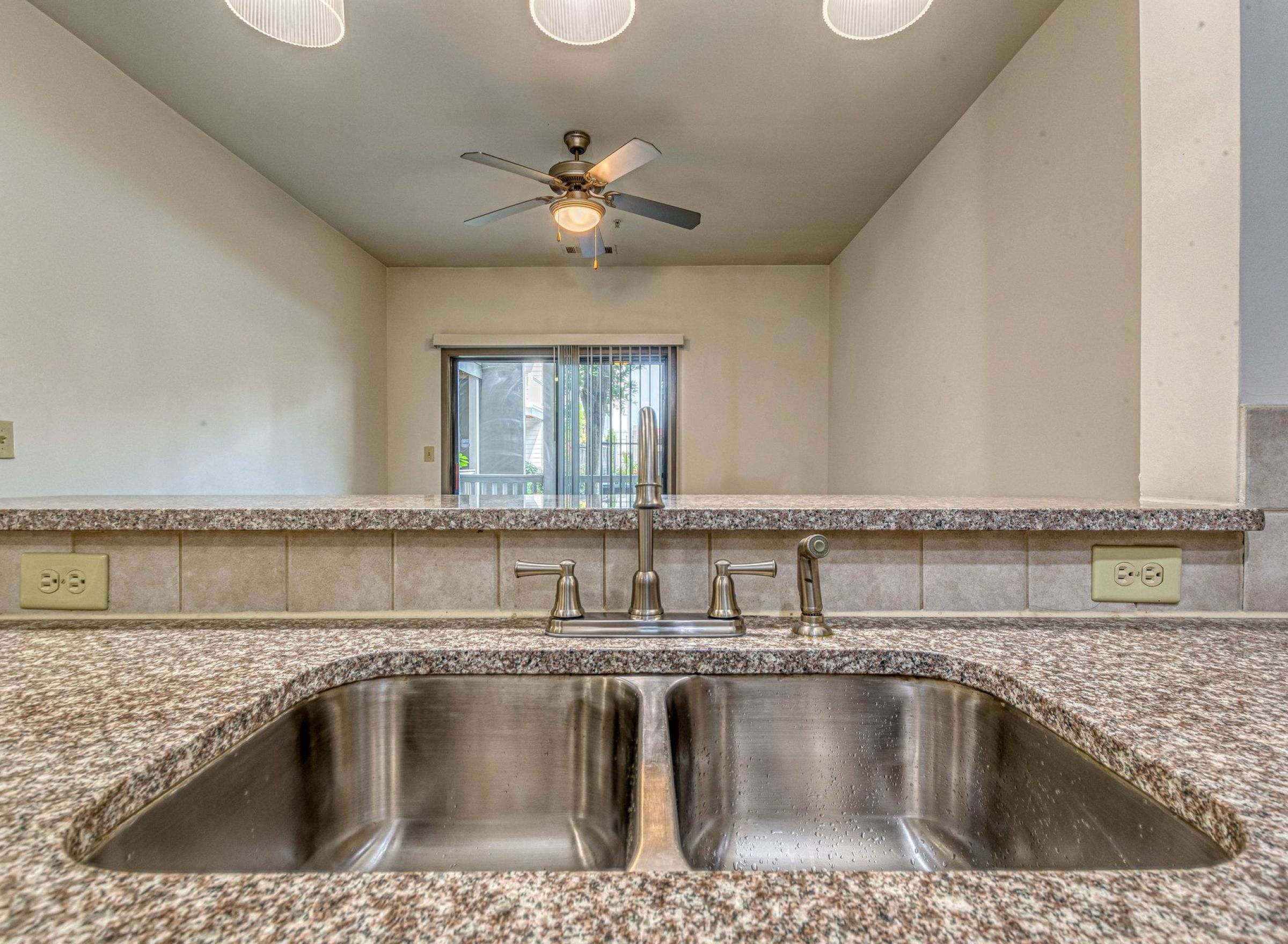 Double kitchen sink with granite countertop, backsplash, and view of ceiling fan and sliding glass door at Apex West Midtown.