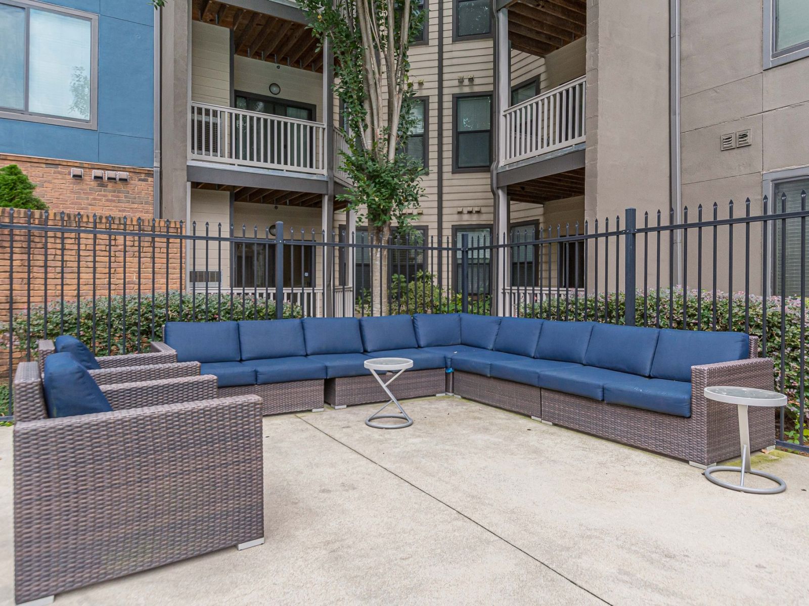 Outdoor patio with a wicker sectional sofa, blue cushions, two side tables, and apartment buildings behind a fence at Apex West Midtown.