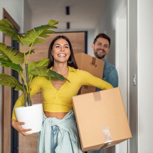 Smiling woman carrying a potted plant and man holding boxes while moving into a home.