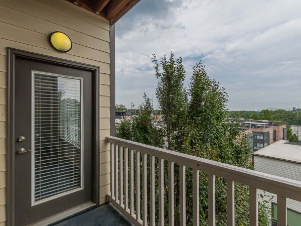 Small balcony with a glass door, railing, and view of trees and city buildings under a cloudy sky at Apex West Midtown.